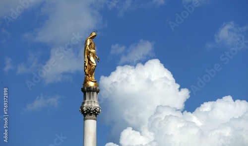 Statue of golden Mother Mary high in the blue sky with big clouds near Cathedral religion background