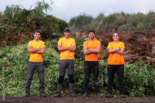Professional team of smiling gardeners and workers standing among piles of pruned branches and green vegetation