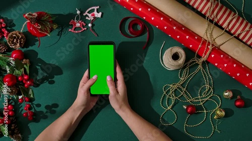 Top view of women hands typing on a smartphone with a green screen against a dark green festive background. The phone is surrounded by festive wrapping materials and Christmas tree ornaments.