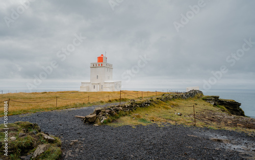 View of a striking white lighthouse with a vibrant orange top stands proudly against the moody sky in Reynisfjara Beach, Myrdalshreppur, Iceland.