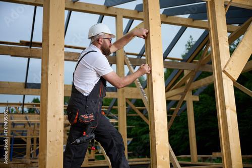 Construction worker measuring wooden beam with tape measure at building frame. Builder in safety helmet working on wooden house construction site outdoors.