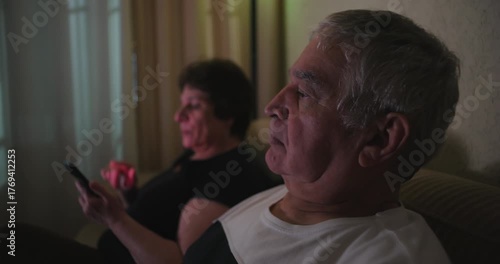Elderly couple seated on a couch, husband focused on TV while wife scrolls through her phone, depicting a peaceful and ordinary living room scene