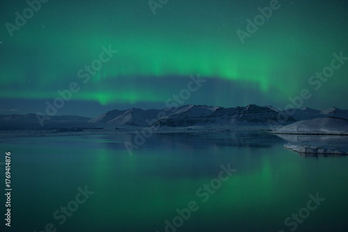 View of the ethereal green aurora borealis dances above the icy lagoon, reflecting in the still waters beneath the snow-capped mountains, Jokulsarlon, Sveitarfélagio Hornafjorour, Iceland.