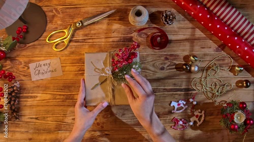 Top view of a woman hands adjusting a gift wrapped in kraft paper, tied with twine, decorated with fir branches and red berries, on a rustic wooden table using scissors and ribbon. Cozy handmade