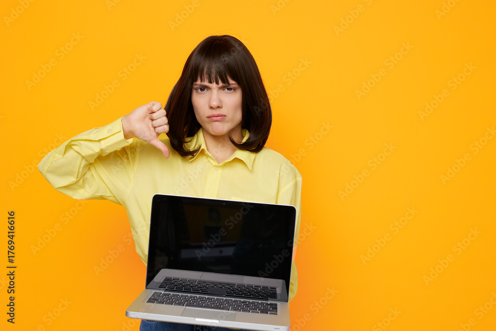 Fototapeta premium Young woman wearing a yellow shirt stands against a vivid orange backdrop, holding a laptop and giving a thumbs down, signaling dissatisfaction, rejection, or negative feedback in a modern tech scene.