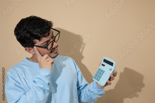 Man with glasses looking at a blue calculator with a thoughtful expression on a light brown background with copy space, concept of financial analysis and economic planning