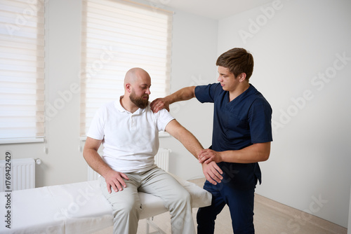Chiropractor adjusting male patient’s shoulder in clinic. Physiotherapist performing manual therapy to relieve pain, improve mobility, support rehabilitation after injury or musculoskeletal disorder.