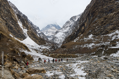 A group of people are trekking a rocky path in the Himalayas toward Annapurna Base Camp, Nepal. Snow dusts the ground and tall mountains surround them.