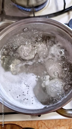 Top down view of water rapidly boiling to cook eggs inside a stainless steel pot. Clear water, white foamy bubbles, and steam rise from the hot liquid on a gas stove in a home kitchen environment.