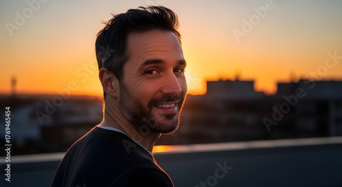 Smiling man looking back over his shoulder on a rooftop at sunset, with warm sunlight and city buildings in the background.