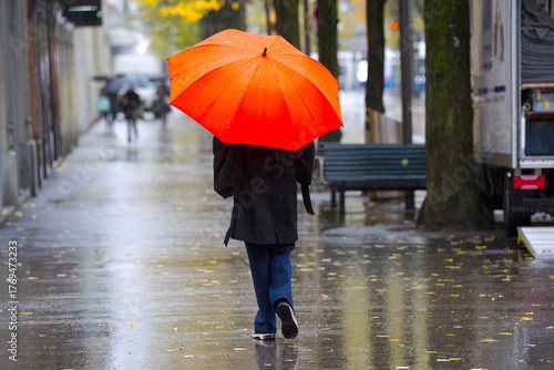 Rear view of young woman with orange umbrella walking on pavement of famous shopping mile named Bahnhofstrasse on a rainy autumn day. Photo taken October 27th, 2025, Zurich, Switzerland.