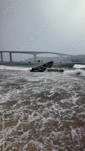 Under a stormy sky, a boat is tossed by rough surf close to the beach, while a large bridge spans the water behind it. ( in slow motion)