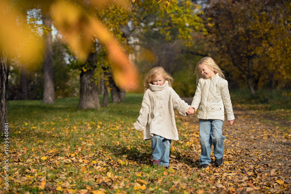 Fototapeta premium Two sisters walking through an autumn park holding hands. Warm natural light and fall colours create a dreamy, emotional atmosphere of childhood and nature.