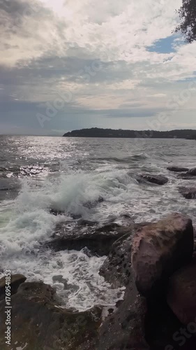 Under a cloudy sky, waves break against a rocky coast, while sunlight shimmers on the sea in front of a distant landmass. ( in slow motion ) 
