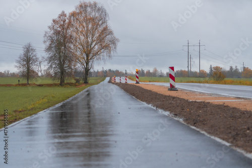 Wet rural road under construction on rainy autumn day, reflective asphalt and safety signs in countryside landscape, transportation and infrastructure concept