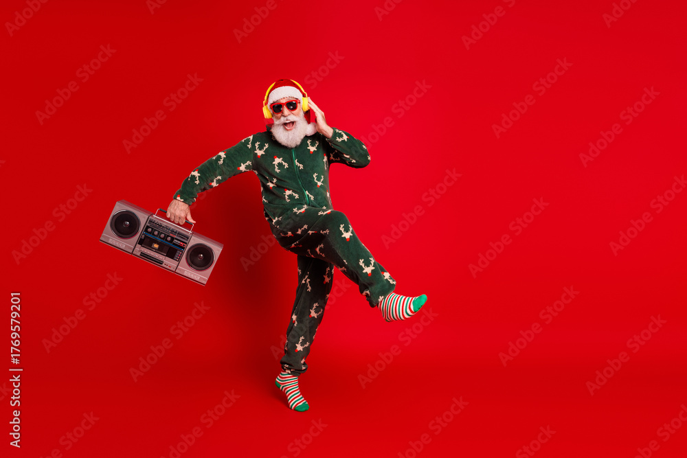 Fototapeta premium Santa in green pajama with a boombox and headphones dances against a red background for a playful Christmas stock photo
