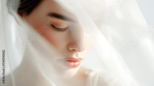 Bride in white tulle veil with closed eyes and natural makeup. Soft focus bridal beauty portrait with delicate features, peaceful expression and ethereal light. Wedding preparation moment concept.