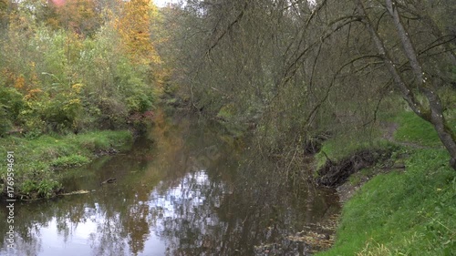 A peaceful river mirrors sky and foliage as a narrow path curves along its bank, framed by drooping branches and early autumn’s golden tones.