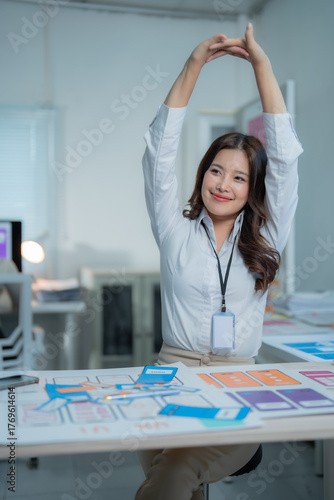 Young Asian woman stretching arms over head at office desk, taking a refreshing break from designing UI UX wireframes, promoting well-being and productivity