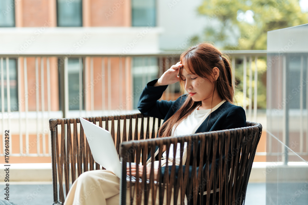 Fototapeta premium Asian businesswoman suffering from severe headache and stress, holding her head while working on a laptop remotely