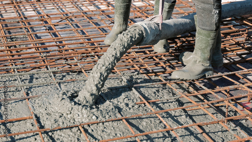 Concrete being poured over steel reinforcement grid. Workers in rubber boots pour wet concrete over a rusty rebar framework during the construction of a reinforced concrete floor.