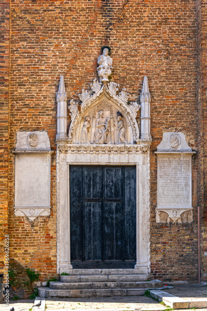 Obraz premium Gothic church entrance with stone relief in Venice. Elaborate Gothic-style church doorway in Venice, with weathered wooden doors and detailed stone religious relief above the entrance.