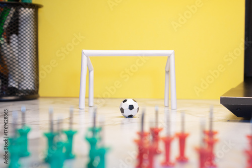 Tiny ball in front of a white goalpost on an office desk with a yellow background and thumbtacks in the foreground simulating soccer teams