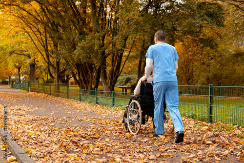 Elderly woman with grey hair sitting in wheelchair during walk in the park in autumn with male nurse in blue uniform helping