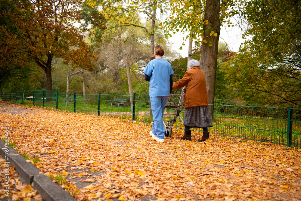 Obraz premium Elderly woman taking a walk through the park in autumn with help of walker and caretaker aid