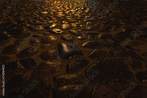 Wet cobblestone street illuminated by warm night lights – close-up texture of old stone pavement after rain, glistening reflections in darkness