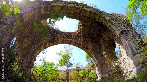 Ancient stone arches overgrown with lush green foliage under a bright sky.