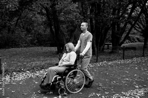 Happy elderly woman with grey hair sitting in wheelchair during walk in park with help of caretaker in blue uniform
