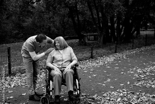 Happy elderly woman with grey hair sitting in wheelchair during walk in park with help of caretaker in blue uniform