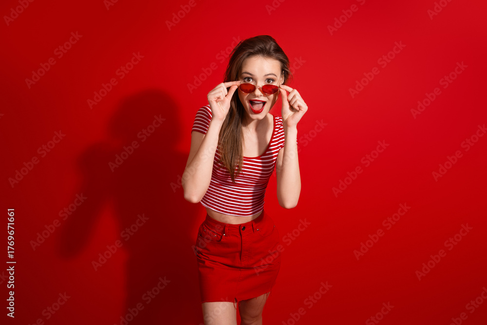 Obraz premium Young stylish woman in red outfit with sunglasses posing on a red background expressing surprise and joy