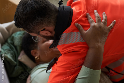 Middle aged Black woman lying on couch being assisted by young adult Caucasian male paramedic wearing uniform, paramedic leaning over providing emergency medical care