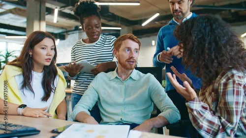 Coworkers debating project results around desk. Man gesturing while explaining graphs on paper as team discusses strategy. Group exchanging ideas and opinions in collaborative office atmosphere.