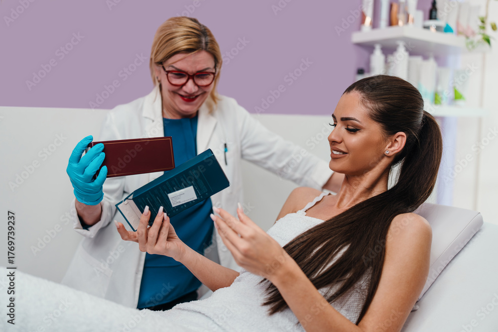 © Dusko - Cosmetologist in blue gloves and lab coat presenting various skincare products to a smiling female client lying on a treatment bed in a modern beauty clinic