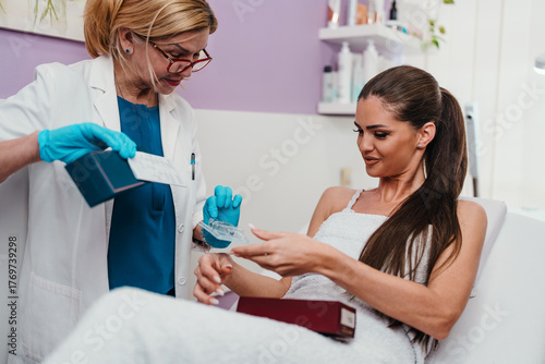 Cosmetologist in blue gloves and lab coat presenting various skincare products to a smiling female client lying on a treatment bed in a modern beauty clinic
