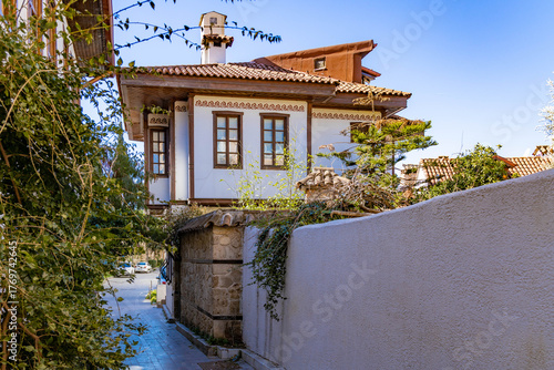 A narrow cobblestone street winds between traditional Ottoman houses with characteristic cumba balconies and sunlit tile roofs in historic Kaleici. Antalya, Turkey.

