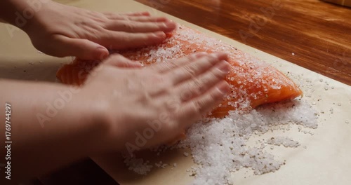 Salting fresh salmon fillet with sea salt on wooden table. Preparing homemade salted salmon, traditional curing process. Close-up of hands salting raw salmon fillet with coarse salt