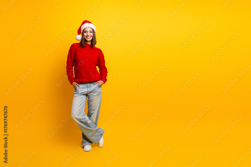 Fototapeta premium Cheerful young woman in a red Christmas sweater and Santa hat posing casually against a bright yellow background for holiday fashion and lifestyle use