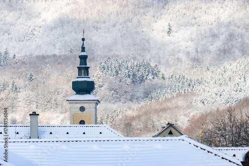 Winter in a mountain village. Snow-covered roofs of buildings and a church tower, with a snowy forest in the background.