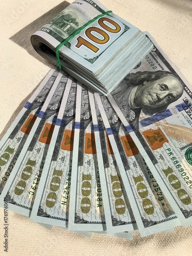 Thick bundle of United States $100 dollar bills secured with a green rubber band, resting on a fan of US $100 dollar bills, symbolizing savings, financial growth, budgeting, and future investment.