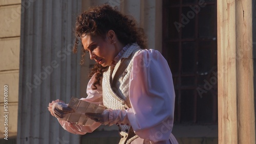 Fotografie An elegant woman in historical clothing stands between the massive, weathered columns of an ancient building and reads a letter in the warm evening light