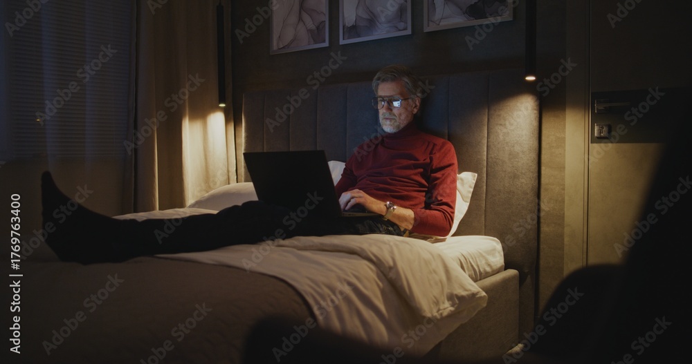 Fototapeta premium Mature Professional Sits on Bed Using Laptop Late at Night in Hotel Room. Man Types on Laptop Keyboard. Concept of Business Traveler Life, Modern Technology, and Late Night Productivity. Handheld.