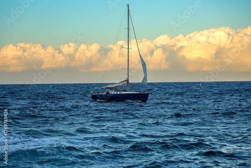 Sleek sailboat gliding across the open sea at sunset, framed by golden clouds and vibrant blue waters. A perfect image of travel, adventure, freedom, and the beauty of sailing.