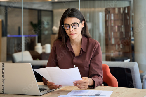 Fotografie Focused latin hispanic business woman working on laptop computer reading financial document trader report in office