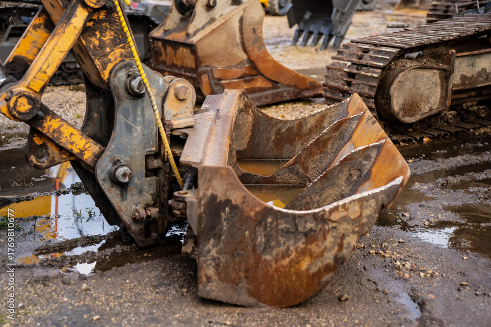 Fototapeta premium Close-up of old excavator bucket attached to hydraulic arm at muddy construction site showing rust, wear and mechanical detail of heavy machinery used for earthmoving and industrial groundworks