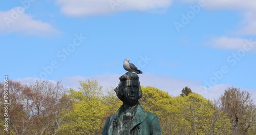 A seagull sits on a weathered bronze statue in a park, leaving white streaks of droppings down the figure's head and coat. Blue spring sky and trees behind it. Urban humor, disrespect, dominance.