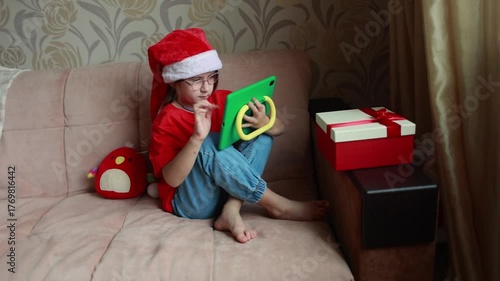 A girl watches a tablet dressed in the Santa hat. New Year's time. A gift for a child.